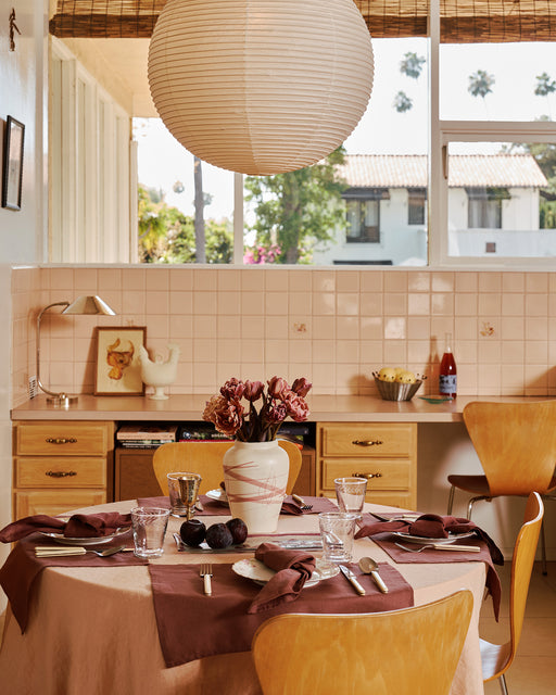 Dining room with a table set for a meal, flowers in the center, and a view of a house from the window. On the table is the 100% French flax tablecloth in terracotta and four napkins and placemats in cacao.