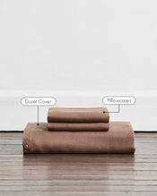 A folded duvet cover in a warm taupe-brown colour called Portobello in 100% french flax linen, sits on a wooden floor against a white wall. Two stacked pillowcases in the same shade sit on top of the duvet. Two callout labels point to the stacked duvet cover and pillowcases, identifying the product components. - view