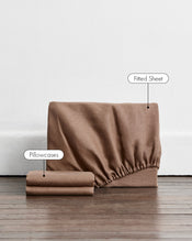A 100% french flax linen fitted sheet and stacked pillowcase covers in a brown shade called portobello. White labels with black text point to the fitted sheet and pillowcases, set against a white wall and dark wood floor. - view