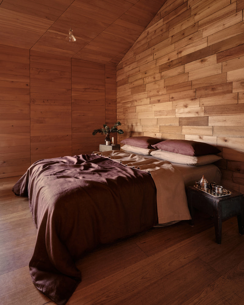 A cozy bedroom with a bed dressed in 100% french flax linen cacao duvet and pillowcases, and fitted and flat sheets in a brown-taupe colour Portobello, set against a wall and ceiling made of warm wood paneling. A small bedside table holds a glass and a small decorative object.