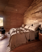 A rustic bedroom with a bed dressed in 100% french flax linen in the shade Portobello, a brown-taupe, set against a wood-paneled wall and matching wood ceiling, with a window and chair visible on the left. - view