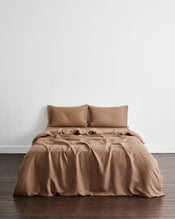 A bed dressed in a draped brown-taupe bedding set in 100% french flax linen with two matching pillows, set against a plain white wall and dark wood flooring. - view