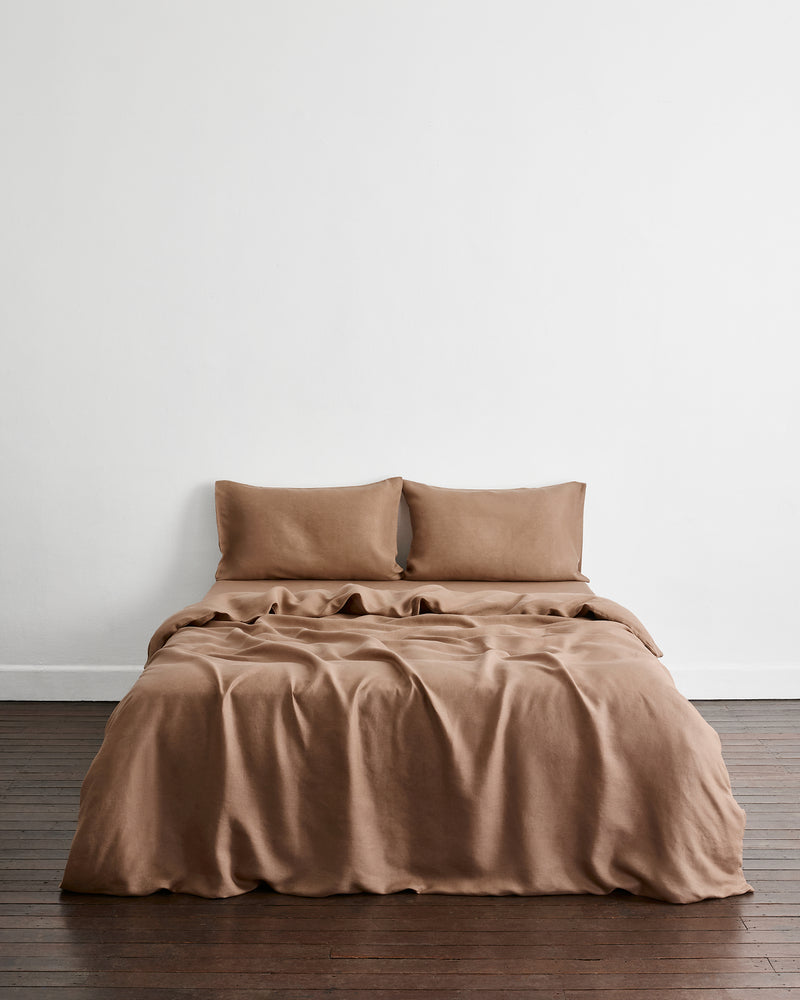 A bed dressed in a draped brown-taupe bedding set in 100% french flax linen with two matching pillows, set against a plain white wall and dark wood flooring.
