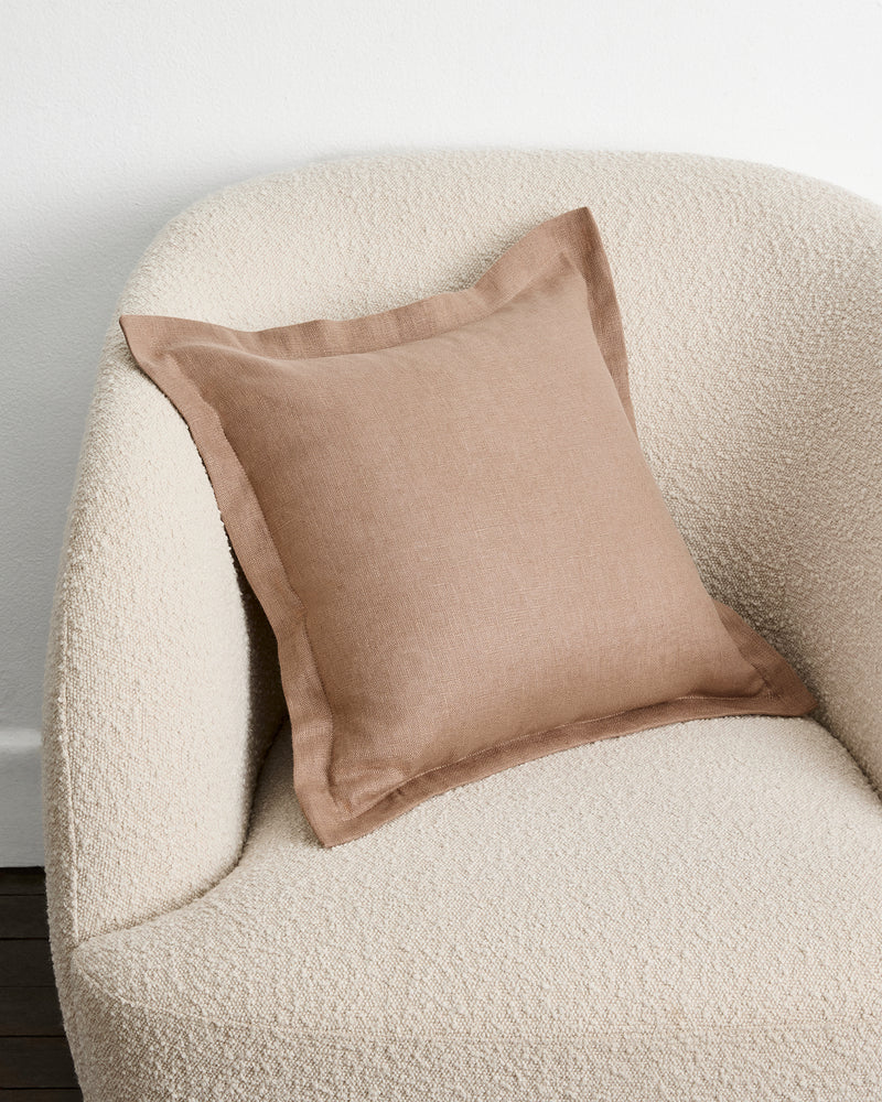 A square warm taupe cushion cover in 100% french flax linen with a subtle border detail, resting on a textured cream boucle armchair against a plain white wall.