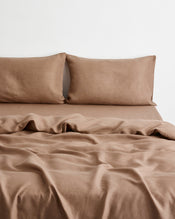 A bed dressed in a soft, draped brown-taupe bedding set in 100% french flax linen with two matching pillowcases against a plain white wall. - view