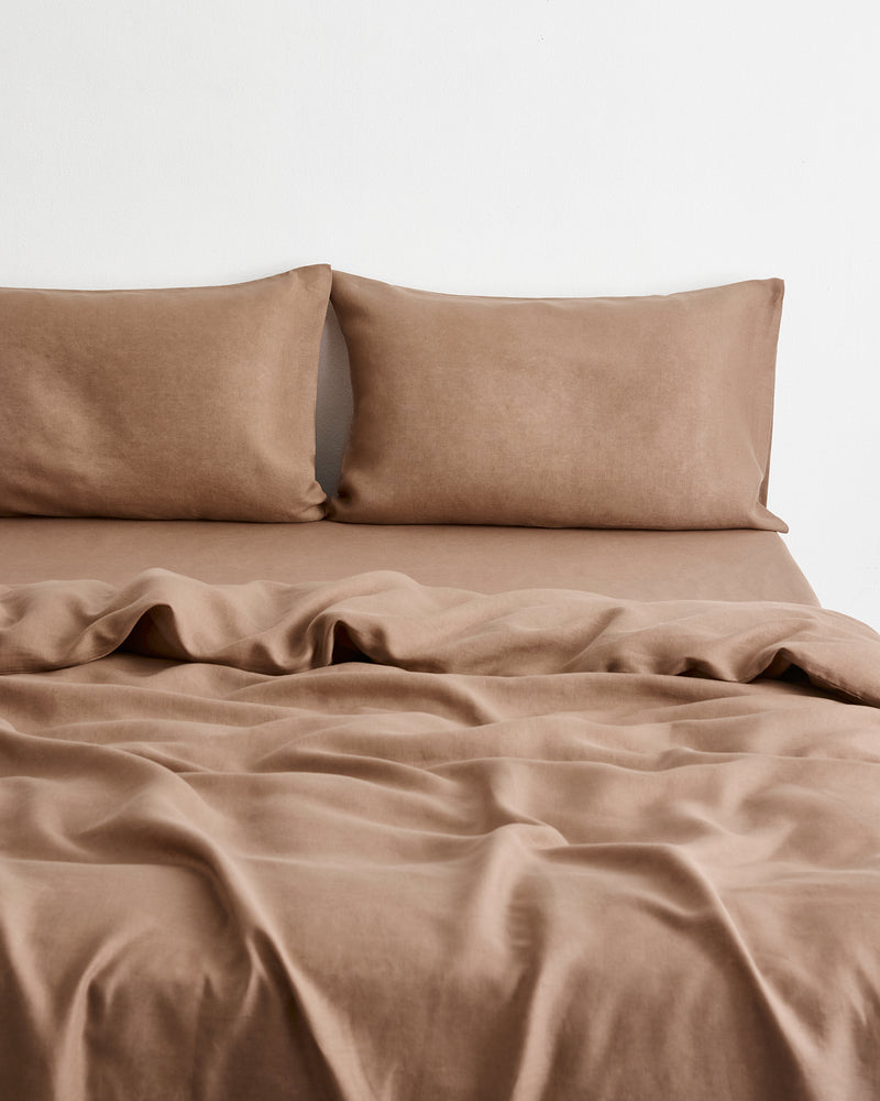 A bed dressed in a soft, draped brown-taupe bedding set in 100% french flax linen with two matching pillowcases against a plain white wall.