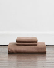 A folded duvet cover in a warm taupe-brown colour called Portobello in 100% french flax linen, sits on a wooden floor against a white wall. Two stacked pillowcases in the same shade sit on top of the duvet.  - view