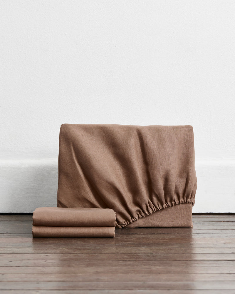 A 100% french flax linen fitted sheet and stacked pillowcase covers in a brown shade called portobello set against a white wall and dark wood floor.

