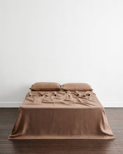 A bed dressed in a smooth, taupe-brown 100% french flax linen sheet and two matching pillows, styled with a ruffled sheet detail on the top, set against a minimalist white wall and dark wood floor. - view