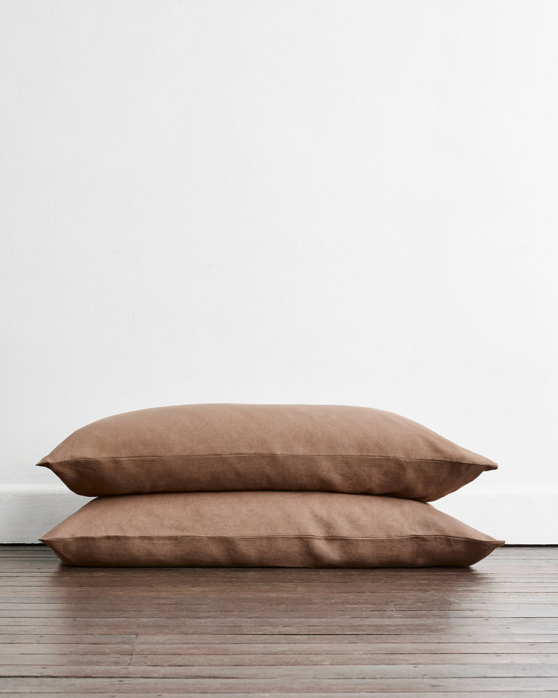 Two king size pillowcases in a brown taupe shade called Portobello available in 100% french flax linen, stacked on top of each other on a dark wood floor against a plain white wall.