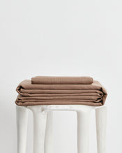 A stack of folded brown 100% french flax linens, including a fitted sheet, flat sheet and pillowcase cover, displayed on a white stool against a plain white background. - view