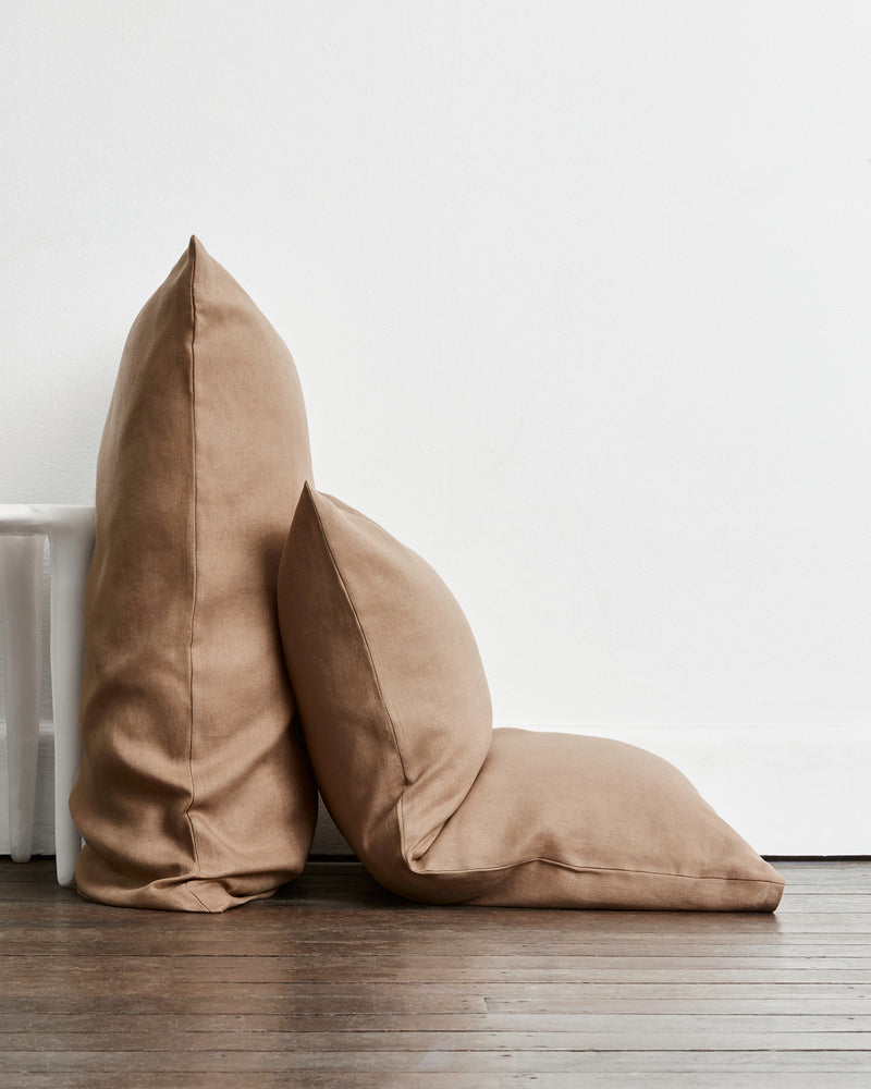 Two brown-taupe standard pillows in 100% french flax linen, one standing upright and one leaning against it, placed on a dark wood floor against a white wall.