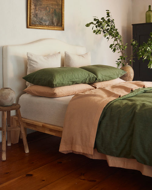 A cozy bedroom with a wooden bed frame, cream headboard, green and beige bedding, and white and green pillows. Plants in vases and rustic decor create a natural, inviting atmosphere.