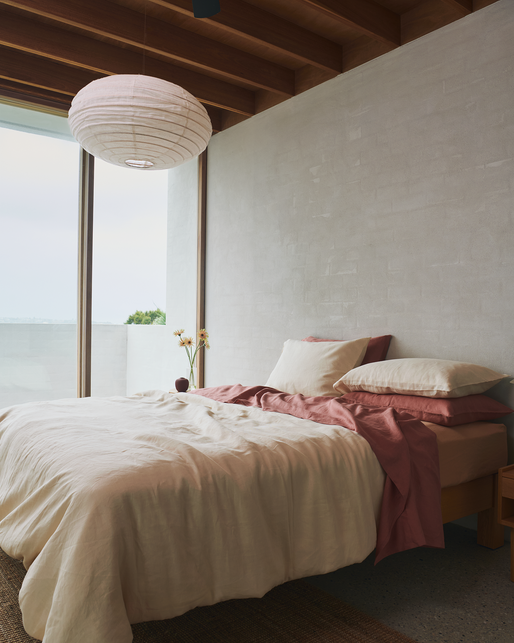 A cozy bedroom with a bed covered in ivory bedding and Bed Threads Pink Clay 100% French Flax Linen Flat Sheet, large windows with natural light, a paper lantern ceiling light, and a vase of flowers on the nightstand.