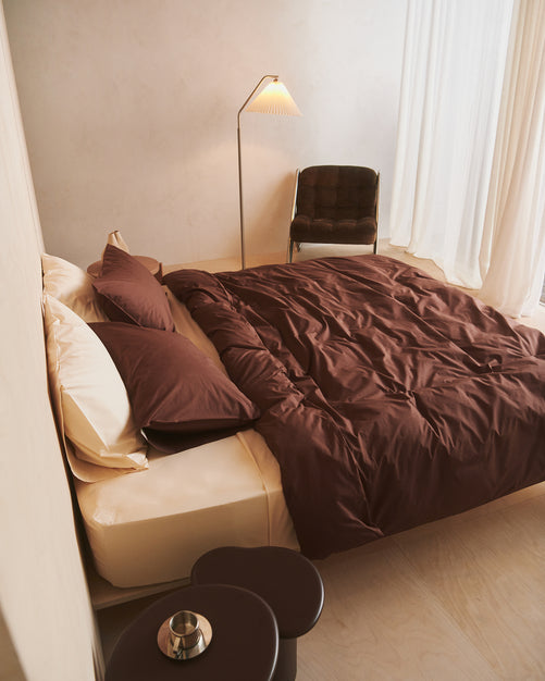 A neatly made bed with deep brown bedding and cream pillows sits beside a modern floor lamp and a dark chair. Light streams in through sheer white curtains, creating a calm, minimalist bedroom setting.