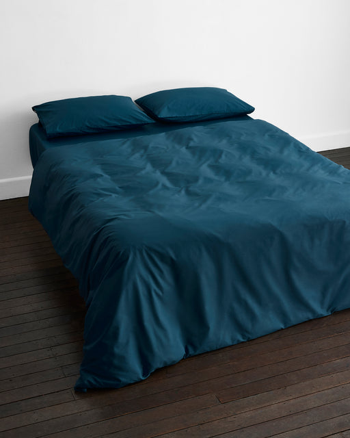 A neatly made bed features the Bed Threads Aegean 100% Organic Cotton Duvet Cover in dark teal with two matching pillows, set against a white wall on a dark wooden floor.