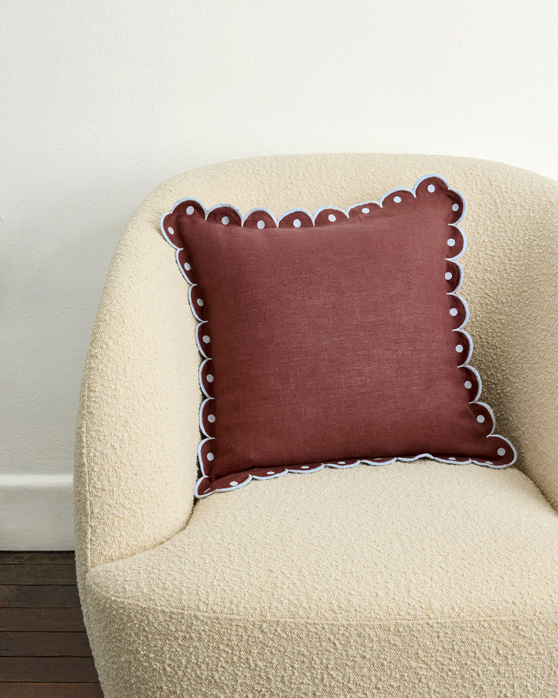 A cream armchair holds a maroon Bed Threads Cacao & Coast 100% French Flax Linen Arch Cushion Cover with scalloped edges and white trim, set against an off-white wall on a dark wooden floor.