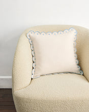The Crème & Coast 100% French Flax Linen Arch Cushion Cover by Bed Threads, cream-colored with embroidered arch border and zip closure, rests on a beige armchair against a white wall and dark wooden floor. - view