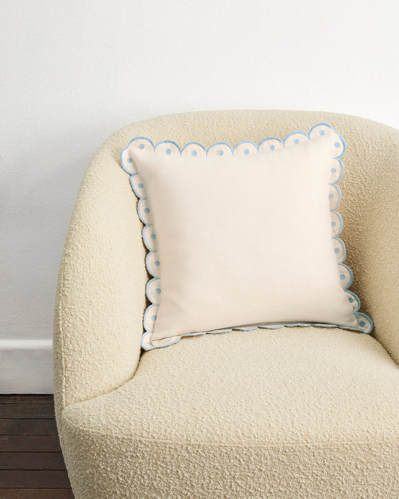 The Crème & Coast 100% French Flax Linen Arch Cushion Cover by Bed Threads, cream-colored with embroidered arch border and zip closure, rests on a beige armchair against a white wall and dark wooden floor.