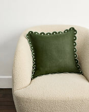 A plush, cream armchair holds the Bed Threads Olive & Sage 100% French Flax Linen Arch Cushion Cover—dark green with scalloped edges and white embroidery—set against a white wall and dark wooden floor. - view