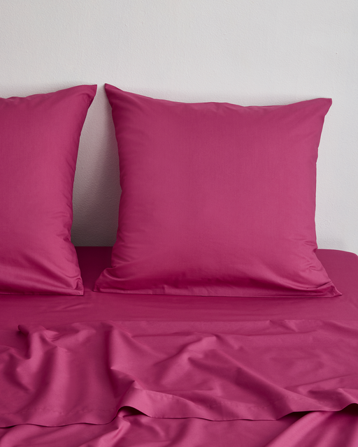 Two Berry 100% Organic Cotton European Pillowcases by Bed Threads rest on a slightly wrinkled magenta bed sheet, set against a plain white wall.