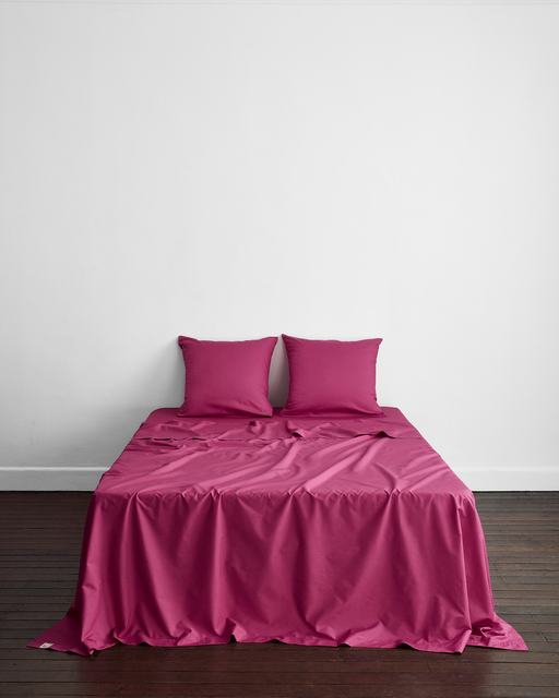 A neatly made bed with magenta Bed Threads Berry 100% Organic Cotton European Pillowcases (Set of Two) stands against a plain white wall on a dark wooden floor.