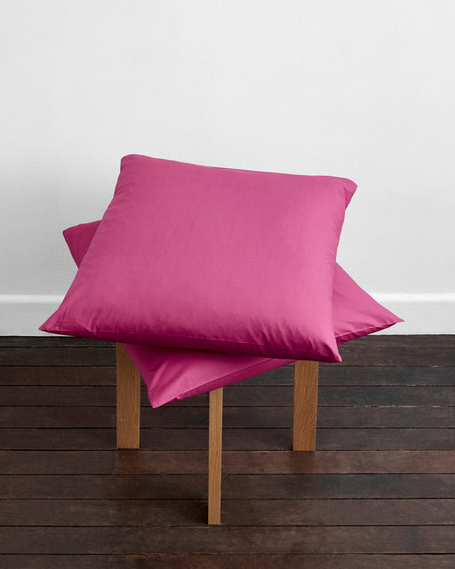 A neatly made bed features Berry Organic Cotton European Pillowcases (Set of Two) by Bed Threads in pink, set against a plain white wall on a dark wooden floor.