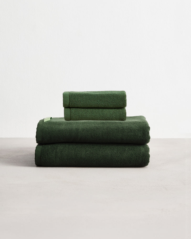 Set of two moss green face washers and two hand towels in 100% cotton terry stacked on a light grey surface with a white background. 