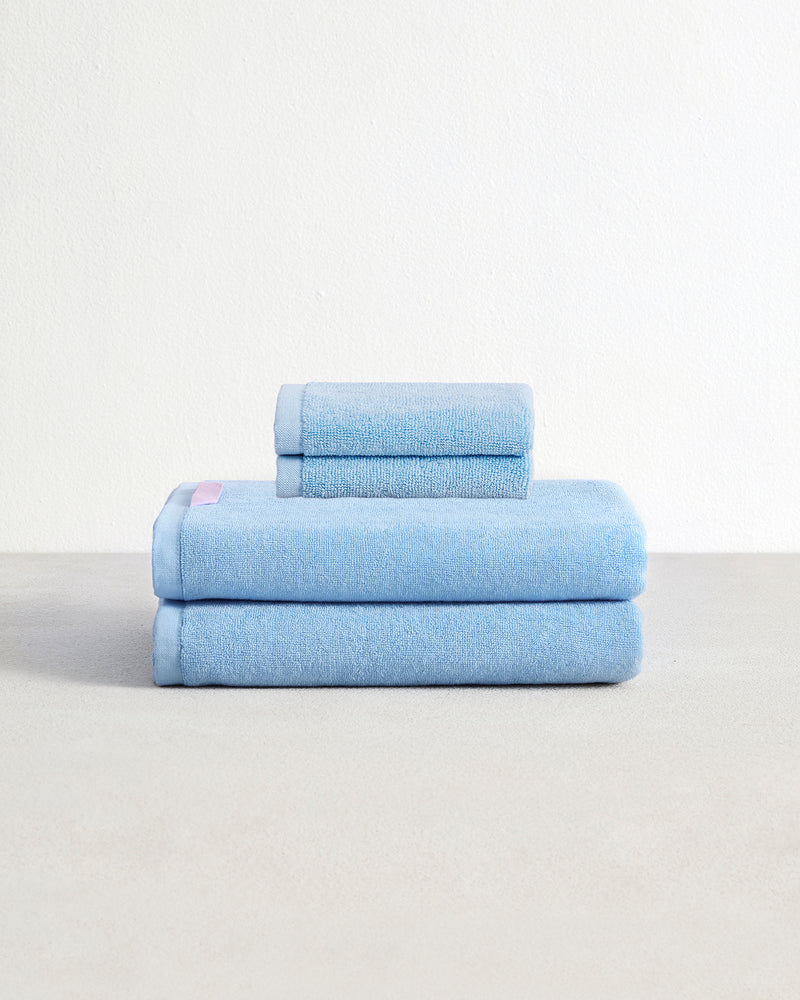 Set of two sky blue face washers and two bath towels in 100% cotton terry stacked on a light grey surface with a white background. 