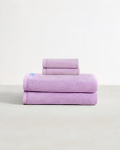 Set of two violet purple face washers and two bath towels in 100% cotton terry stacked on a light grey surface with a white background.  - view