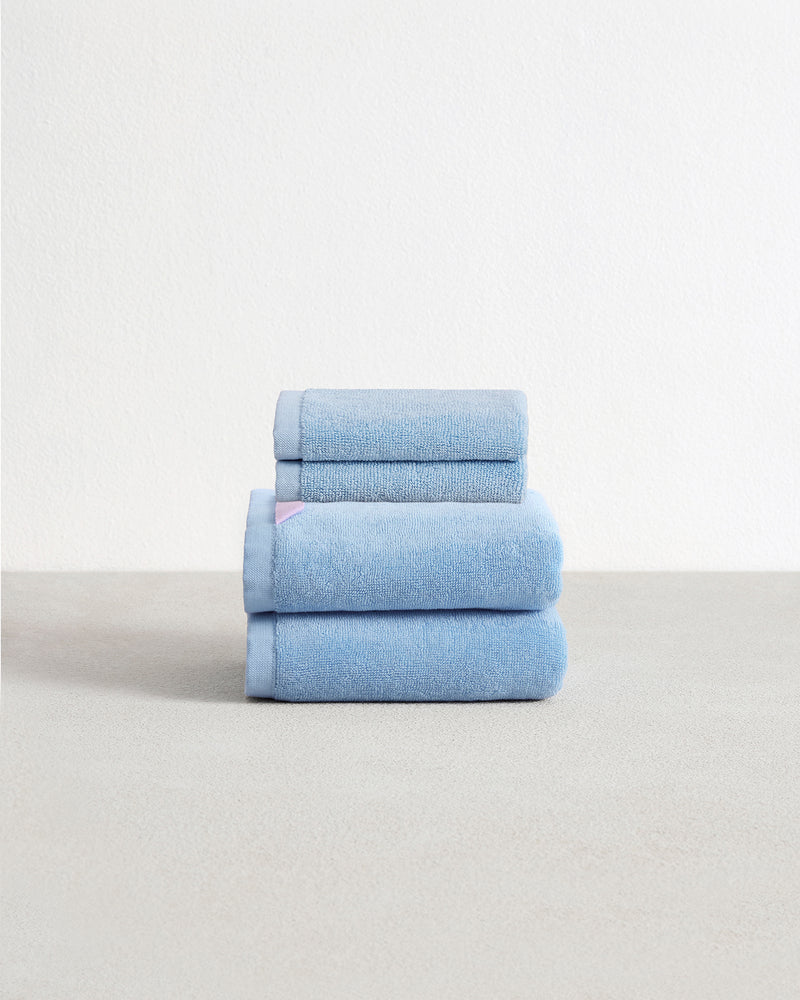 Set of two sky blue face washers and two hand towels in 100% cotton terry stacked on a light grey surface with a white background. 