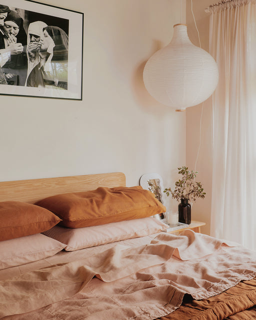 A cozy bedroom with a wooden headboard, Bed Threads’ Rust & Terracotta 100% French Flax Linen Bedding Bundle, a round paper lantern, sheer curtains, a small nightstand with a vase of greenery, and a black-and-white photo on the wall.