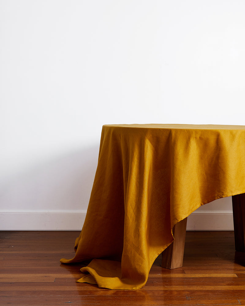 A round wooden table is elegantly covered with the Bed Threads Turmeric 100% French Flax Linen Tablecloth, its rich yellow fabric cascading onto a polished wooden floor set against a plain white wall.