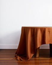 A wooden dining table is adorned with the Bed Threads Rust 100% French Flax Linen Tablecloth, elegantly draped and spilling onto the hardwood floor, set against a plain white wall. - view
