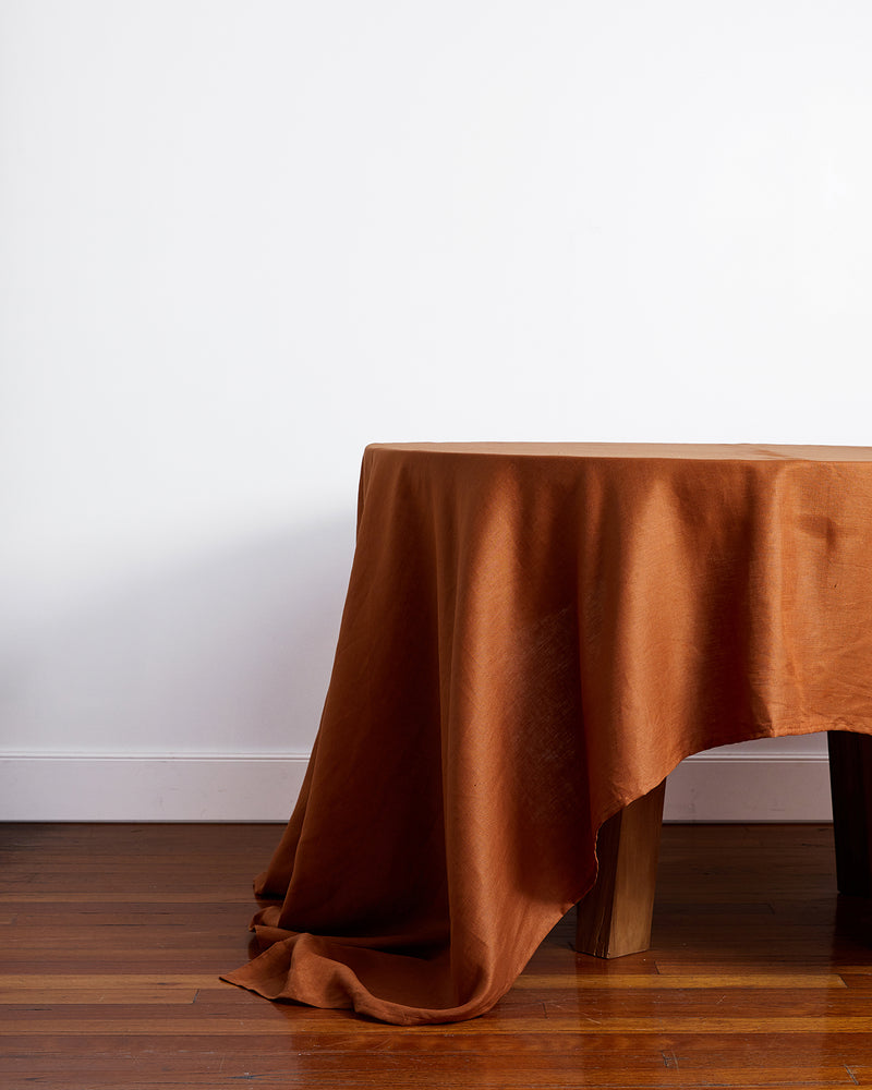 A wooden dining table is adorned with the Bed Threads Rust 100% French Flax Linen Tablecloth, elegantly draped and spilling onto the hardwood floor, set against a plain white wall.