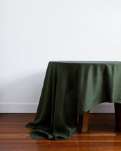 A table featuring the Bed Threads Olive 100% French Flax Linen Tablecloth, its rich green fabric partially concealing the wooden legs, stands on a polished wood floor against a plain white wall. - view