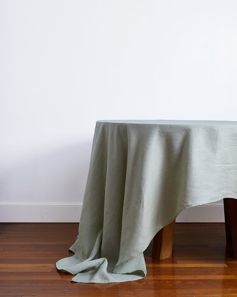 The Bed Threads Sage 100% French Flax Linen Tablecloth in light gray drapes over a round table, pooling onto wood flooring. A plain white wall and baseboard complete this elegant linen table setting.