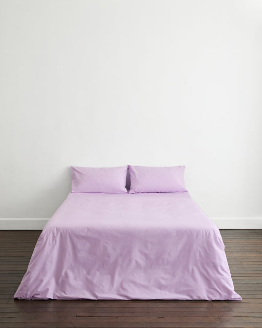 A neatly made bed features the Bed Threads Violet 100% Organic Cotton Duvet Cover and two matching pillows, set against a plain white wall on a dark wooden floor.