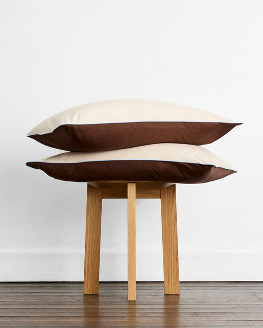 Two pillows with Bed Threads’ Espresso, Vanilla & Sky Piped 100% Organic Cotton Pillowcases (Set of Two) are stacked on a wooden stool against a white wall and wooden floor.