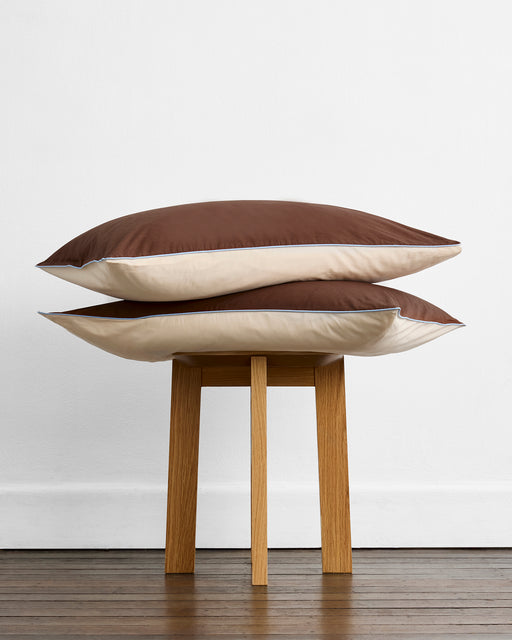 Two Bed Threads Espresso, Vanilla & Sky Piped 100% Organic Cotton Pillowcases are stacked on a wooden stool against a white wall and floor. Each pillowcase features brown and beige tones for a stylish, natural look.