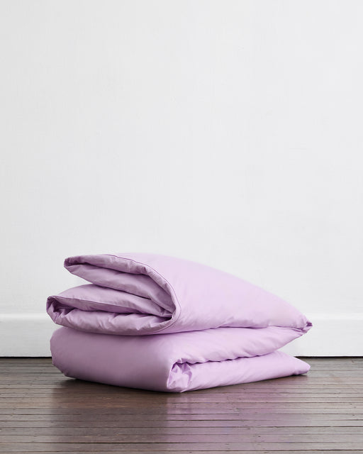 A neatly folded Violet 100% Organic Cotton Duvet Cover by Bed Threads rests on a wooden floor against a plain white wall.