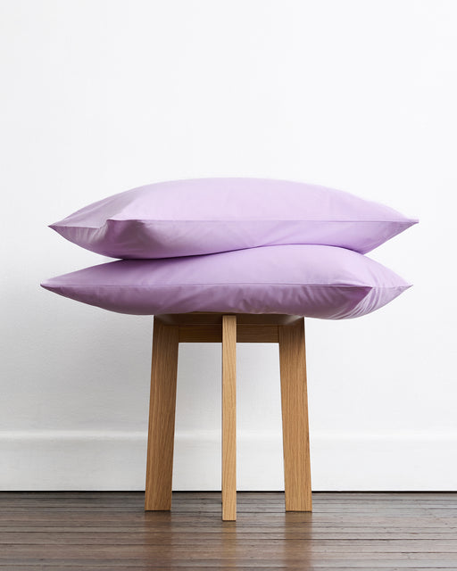Two light purple pillows from the Sky & Violet 100% Organic Cotton Bedding Bundle by Bed Threads, with a percale weave, are stacked on a wooden stool against a white wall and wooden floor.