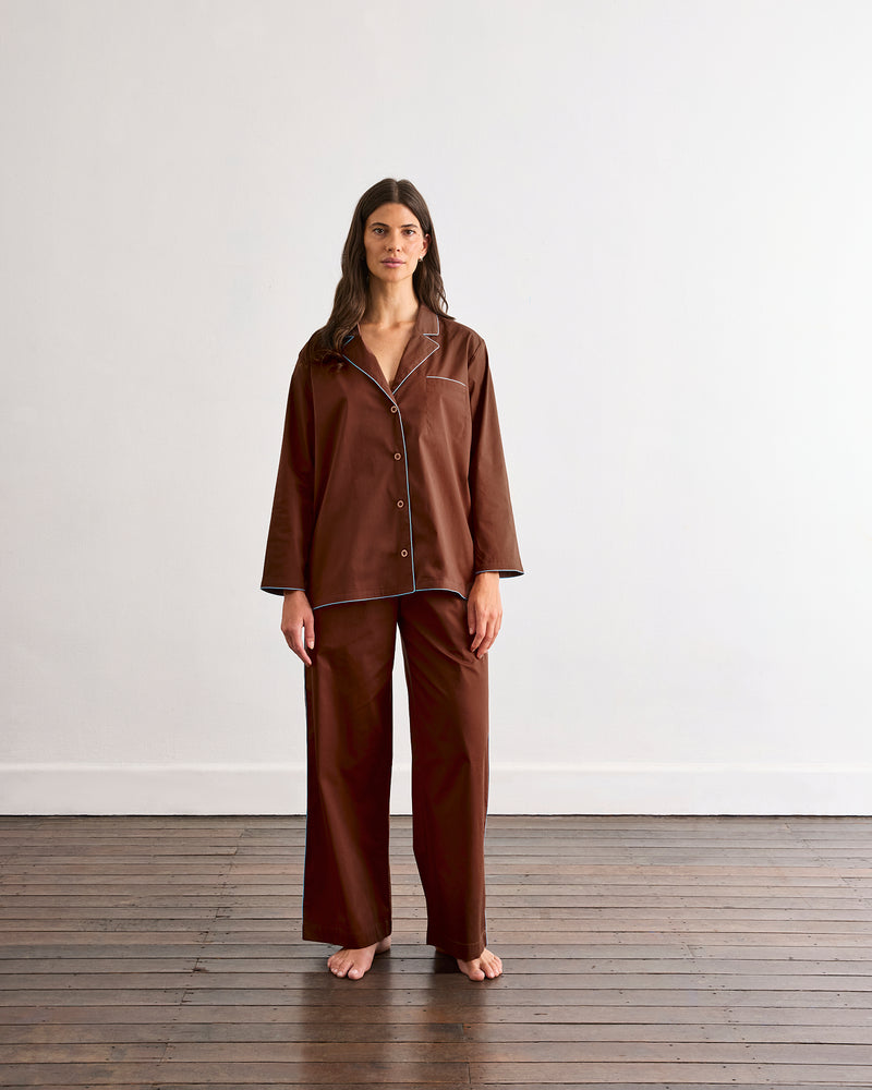 A woman stands barefoot on a wooden floor in front of a plain white wall, wearing the Bed Threads Espresso & Sky Piped Long Sleep Set.