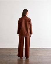 A woman stands barefoot on a wooden floor facing away, wearing the Bed Threads Espresso & Sky Piped Long Sleep Set—loose-fitting pants and matching long-sleeve top—against a plain white wall. - view