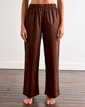 A person stands barefoot on a wooden floor, wearing the Bed Threads Espresso & Sky Piped Long Sleep Set—loose-fitting pants with an elastic waistband and side pockets—against a plain white wall. - view
