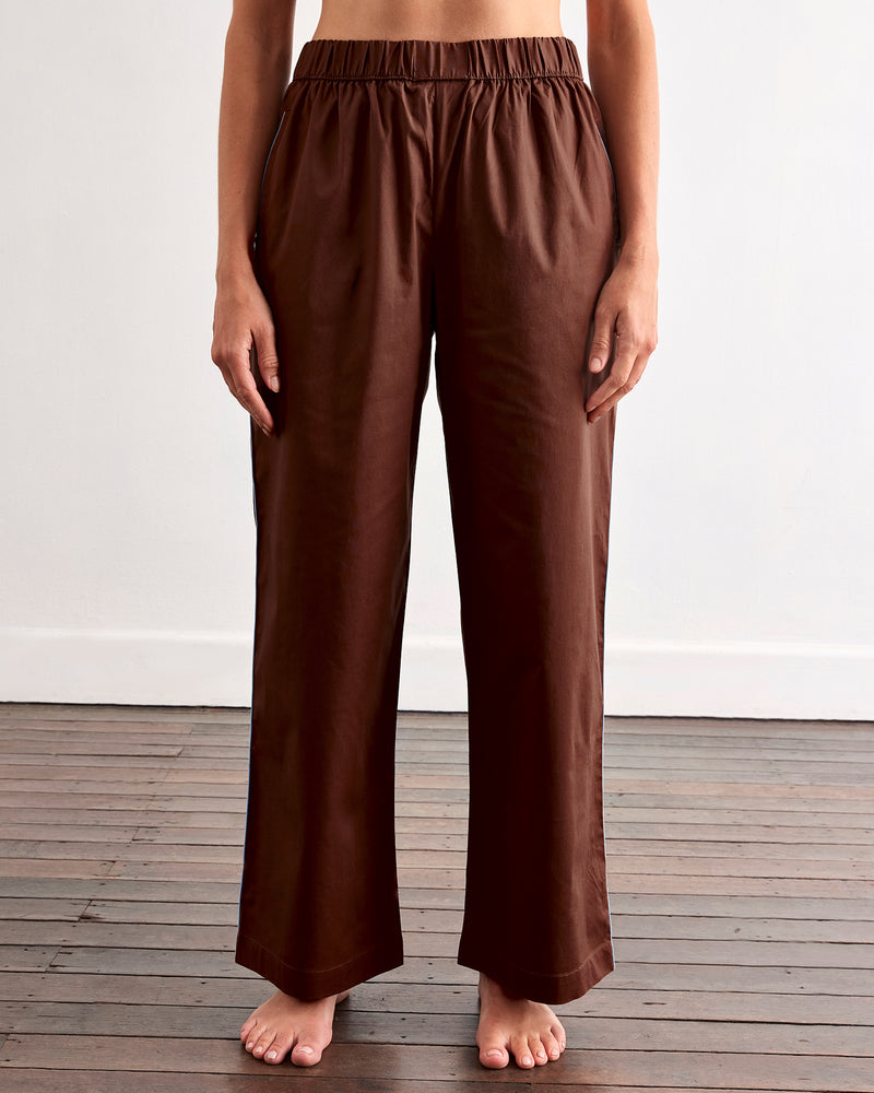 A person stands barefoot on a wooden floor, wearing the Bed Threads Espresso & Sky Piped Long Sleep Set—loose-fitting pants with an elastic waistband and side pockets—against a plain white wall.