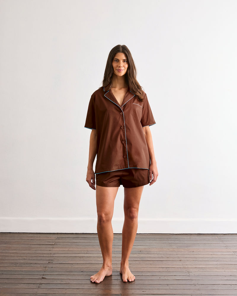 A woman stands barefoot on a wooden floor against a white wall, wearing the Bed Threads Espresso & Sky Piped Short Sleep Set—a lightweight cotton set with a short sleeve shirt and piped shorts featuring crisp white piping.