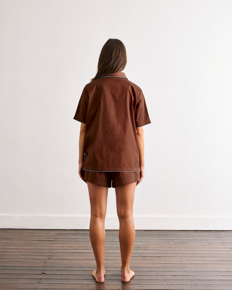 A woman with long brown hair stands barefoot on a wooden floor, facing a white wall. She is wearing the Bed Threads Espresso & Sky Piped Short Sleep Set, featuring a brown short sleeve shirt and matching piped shorts.