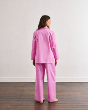 A woman stands barefoot on a wooden floor, facing away from the camera, wearing the Bubblegum & Berry Piped Long Sleep Set by Bed Threads—a loose-fitting, long-sleeved pink pyjama set—against a plain white wall. - view