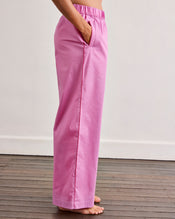 A person stands barefoot on a wooden floor, wearing Bed Threads’ Bubblegum & Berry Piped Pants in light pink cotton with an elasticated waist and side pockets, shown from the side to highlight their comfortable, relaxed fit. - view
